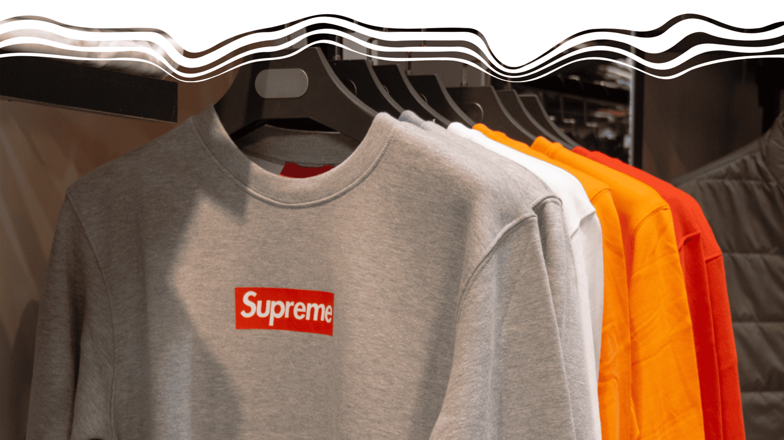 A selection of Supreme sweatshirts hanging on a clothing rack, featuring a standout gray hoodie with the brand’s signature red and white logo. The image showcases various sweatshirt colors, including white, orange, and red, emphasizing custom apparel as a versatile branding tool.